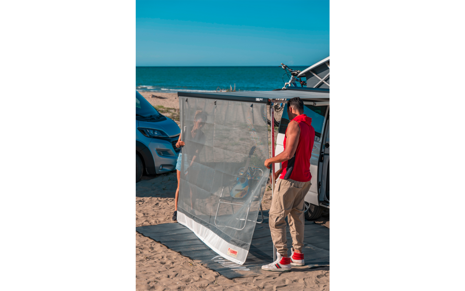 Mann hält ein durchsichtiges Netz vor geöffnetem Camper‑Van am Sandstrand, Meer im Hintergrund, Fahrrad auf dem Dach, Campingstühle sichtbar