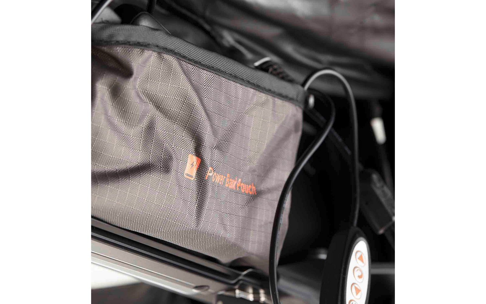 Grau strukturierte Stofftasche mit orangefarbenem Logo und Text 'Power Back Couch', an einem Metallrahmen befestigt; ein schwarzes Kabel verläuft daneben; ein Rad mit weißer Nabe und orangefarbener Markierung sichtbar