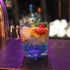 Glas mit einem blauen Cocktail, Eiswürfeln, einer Limettenscheibe, einem Minzzweig und einer Erdbeere auf einer Bar