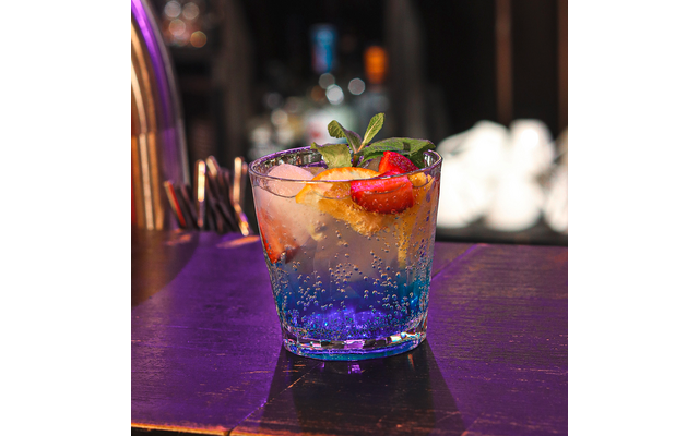 Glas mit einem blauen Cocktail, Eiswürfeln, einer Limettenscheibe, einem Minzzweig und einer Erdbeere auf einer Bar