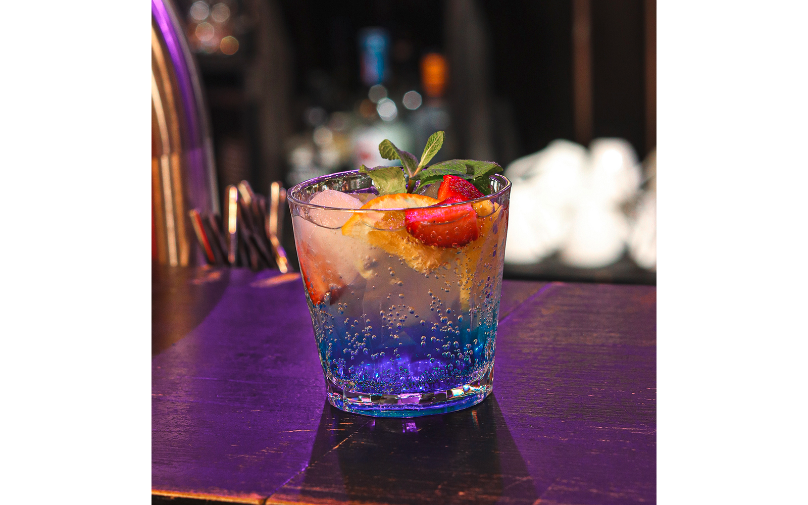 Glas mit einem blauen Cocktail, Eiswürfeln, einer Limettenscheibe, einem Minzzweig und einer Erdbeere auf einer Bar