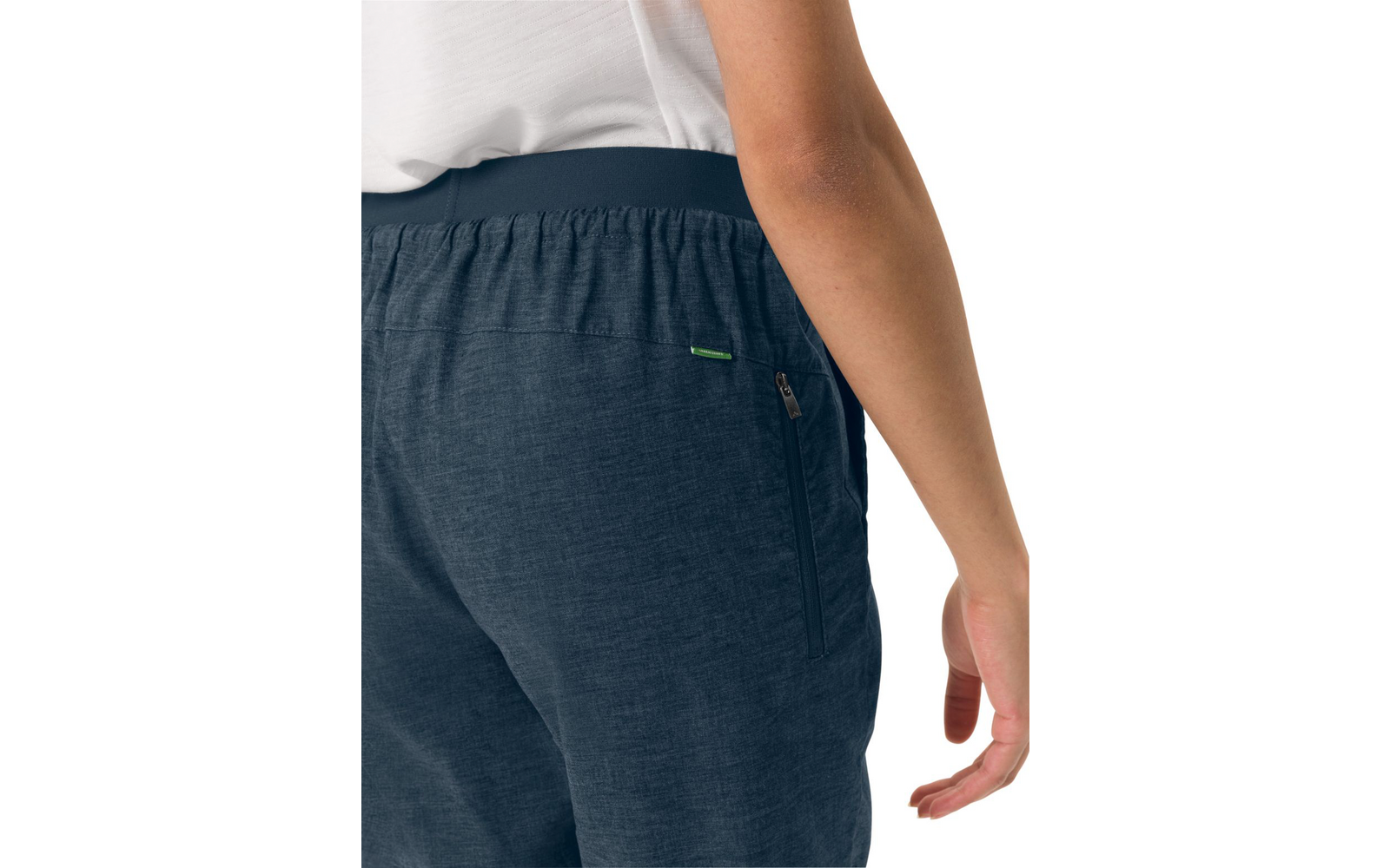 Hip area of a person wearing dark blue pants with an elastic waistband, a side zip pocket and a small green tag; white top and right arm visible