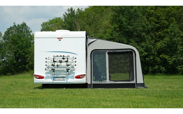 Weißes Wohnmobil von hinten mit Fahrradträger und grauem Vorzelt auf grüner Wiese; Bäume und bewölkter Himmel im Hintergrund