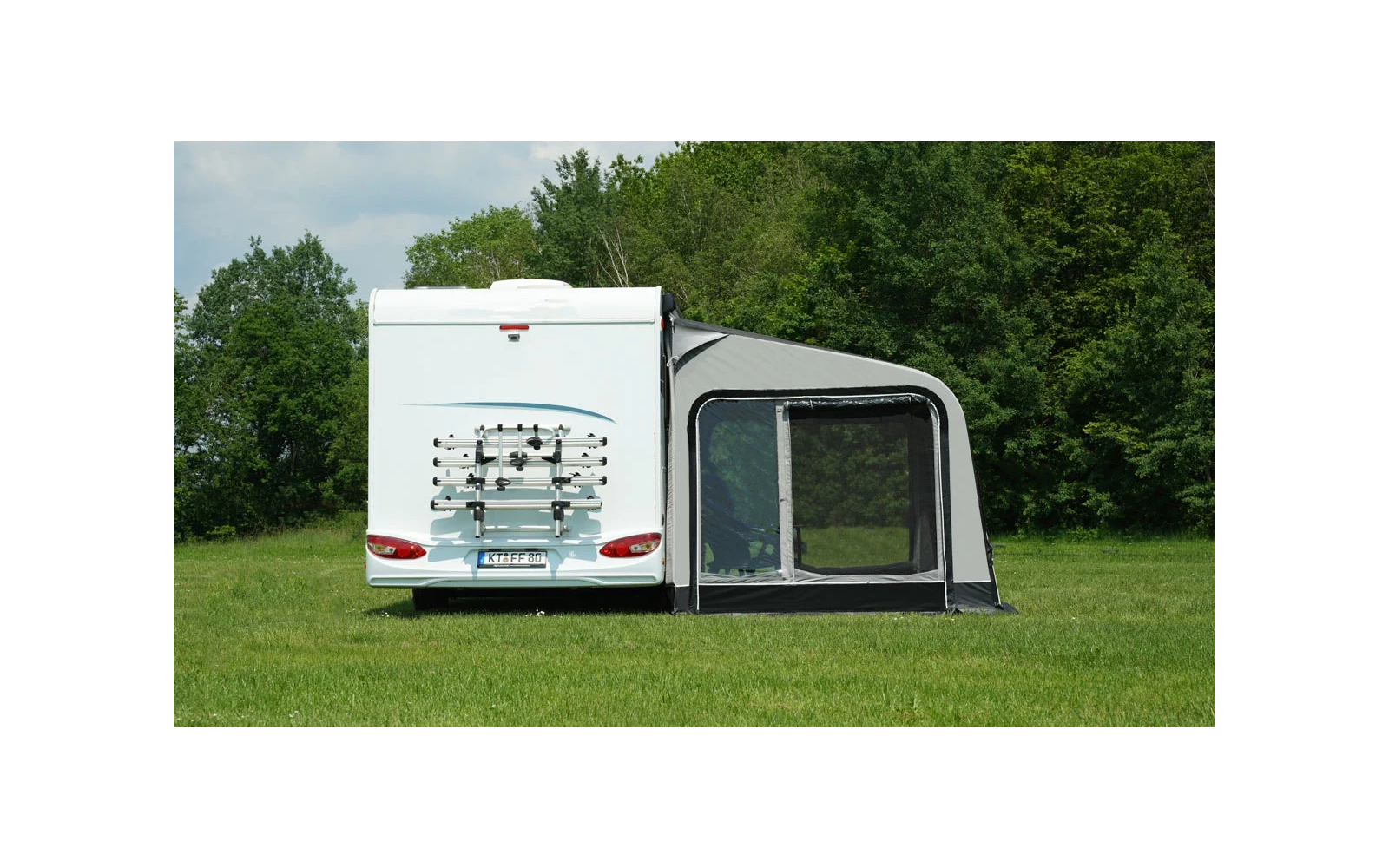 Weißes Wohnmobil von hinten mit Fahrradträger und grauem Vorzelt auf grüner Wiese; Bäume und bewölkter Himmel im Hintergrund