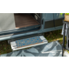 Marchepied métallique déployé d'un camping-car avec tapis de marche bleu et orange à motifs, tapis extérieur gris à motif de feuilles sur l'herbe, porte coulissante ouverte et plancher en bois visible ; bord droit avec pied de table et sac de plante