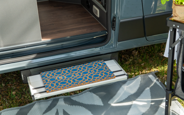 Marchepied métallique déployé d'un camping-car avec tapis de marche bleu et orange à motifs, tapis extérieur gris à motif de feuilles sur l'herbe, porte coulissante ouverte et plancher en bois visible ; bord droit avec pied de table et sac de plante