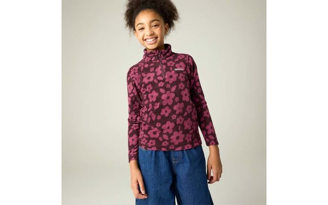 Mädchen steht vor hellem Hintergrund, lächelt, trägt lila-pinkes Langarmshirt mit Blumenmuster und kurzem Reißverschluss sowie blaue weite Hosen; Hände an den Seiten