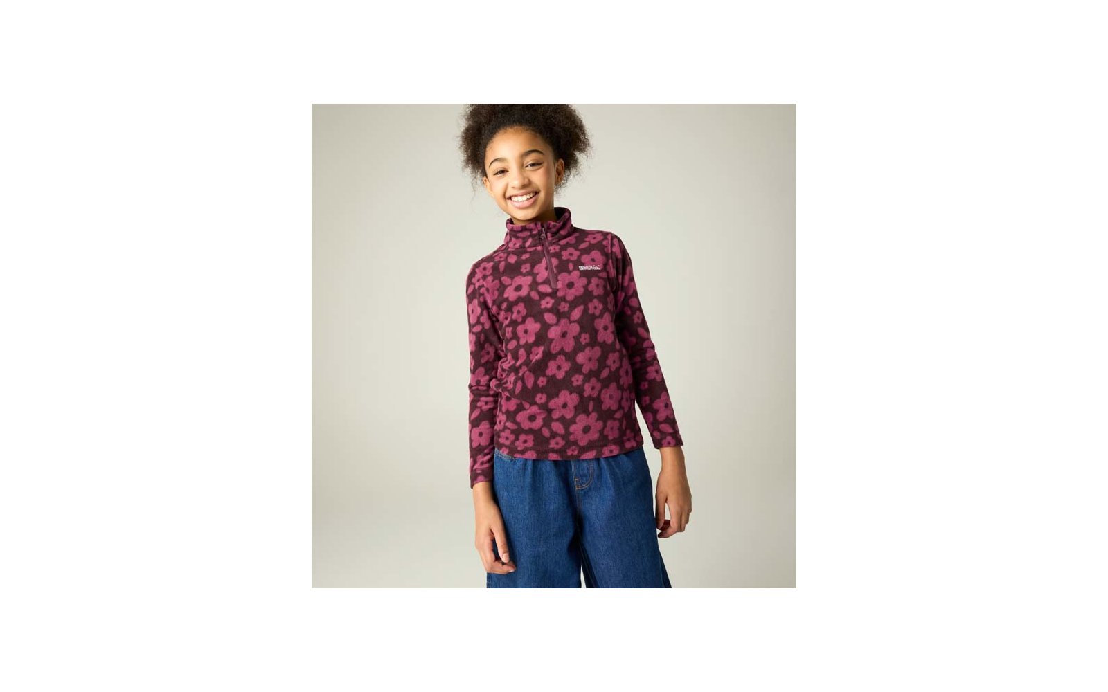 Mädchen steht vor hellem Hintergrund, lächelt, trägt lila-pinkes Langarmshirt mit Blumenmuster und kurzem Reißverschluss sowie blaue weite Hosen; Hände an den Seiten