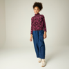Lächelnde Person steht, trägt violetten Rollkragenpullover mit Blumenmuster, weite blaue Jeans und beige Sneaker, heller Hintergrund