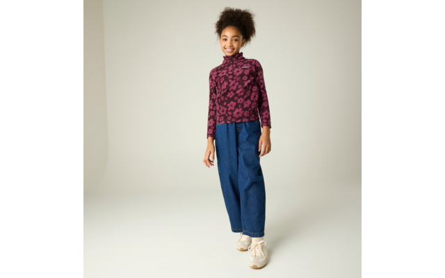 Lächelnde Person steht, trägt violetten Rollkragenpullover mit Blumenmuster, weite blaue Jeans und beige Sneaker, heller Hintergrund