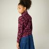 Person mit hochgestecktem lockigem Haar, trägt violett-rosa floral gemustertes Langarmoberteil und blauen Jeansrock, schaut über die Schulter, heller einfarbiger Hintergrund