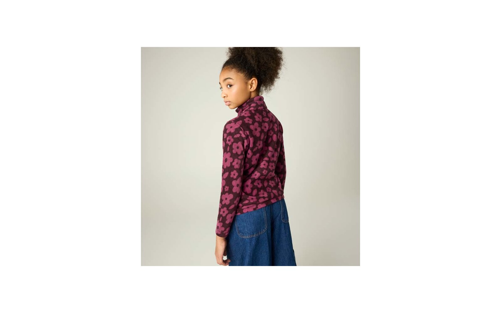 Person mit hochgestecktem lockigem Haar, trägt violett-rosa floral gemustertes Langarmoberteil und blauen Jeansrock, schaut über die Schulter, heller einfarbiger Hintergrund