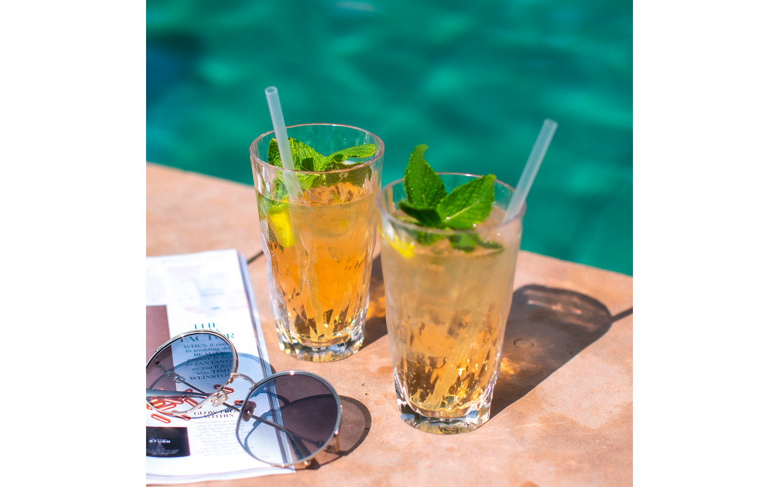 Zwei Gläser mit Eistee und Minzblättern auf einem Tisch neben einer Sonnenbrille am Pool