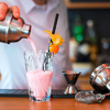 Ein Glas mit einem rosa Getränk und einem Orangendekorationsstück, im Hintergrund eine Hand mit einem Cocktailshaker