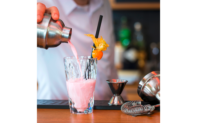 Ein Glas mit einem rosa Getränk und einem Orangendekorationsstück, im Hintergrund eine Hand mit einem Cocktailshaker