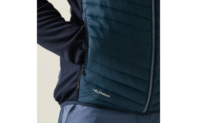 Nahaufnahme des Oberkörpers einer Person mit dunklem Ärmel, gesteppter dunkelblau-grüner Reißverschlussweste mit seitlicher Tasche und kleinem weißen Logo am Saum sowie hellblauer Hose