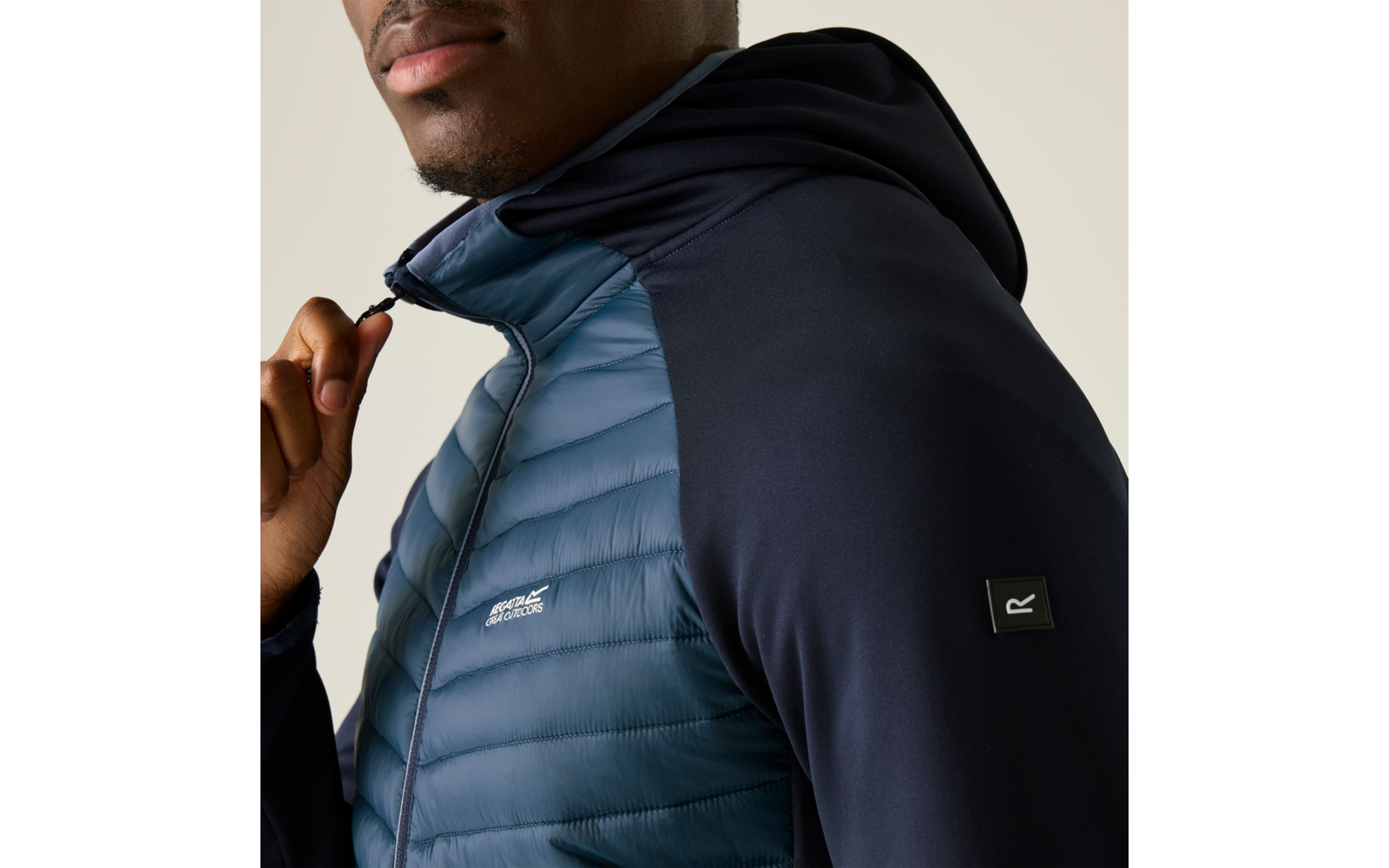 Halbnahes Foto einer Person mit sichtbarem Kinn und Lippen, sie trägt eine dunkelblaue gesteppte Kapuzenjacke und hält mit der Hand den Reißverschluss; schwarze Ärmel mit kleinem quadratischem Patch