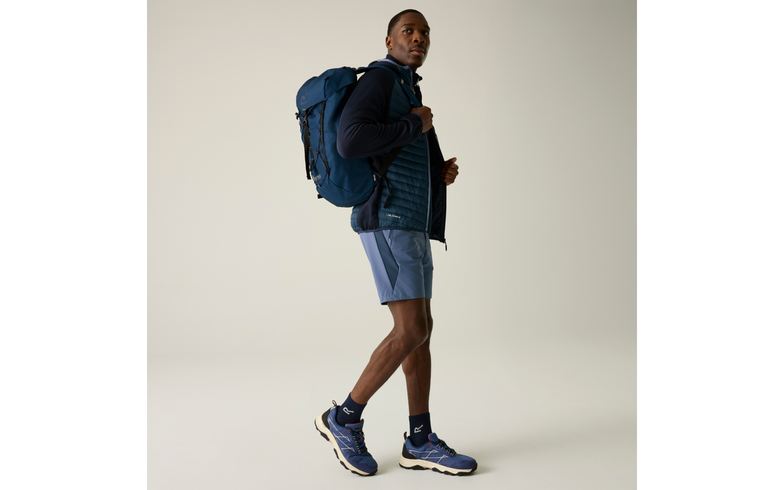 Person in blauer Kleidung mit Rucksack, trägt Shorts, Jacke und Sportschuhe, steht auf hellem Hintergrund und schaut seitlich