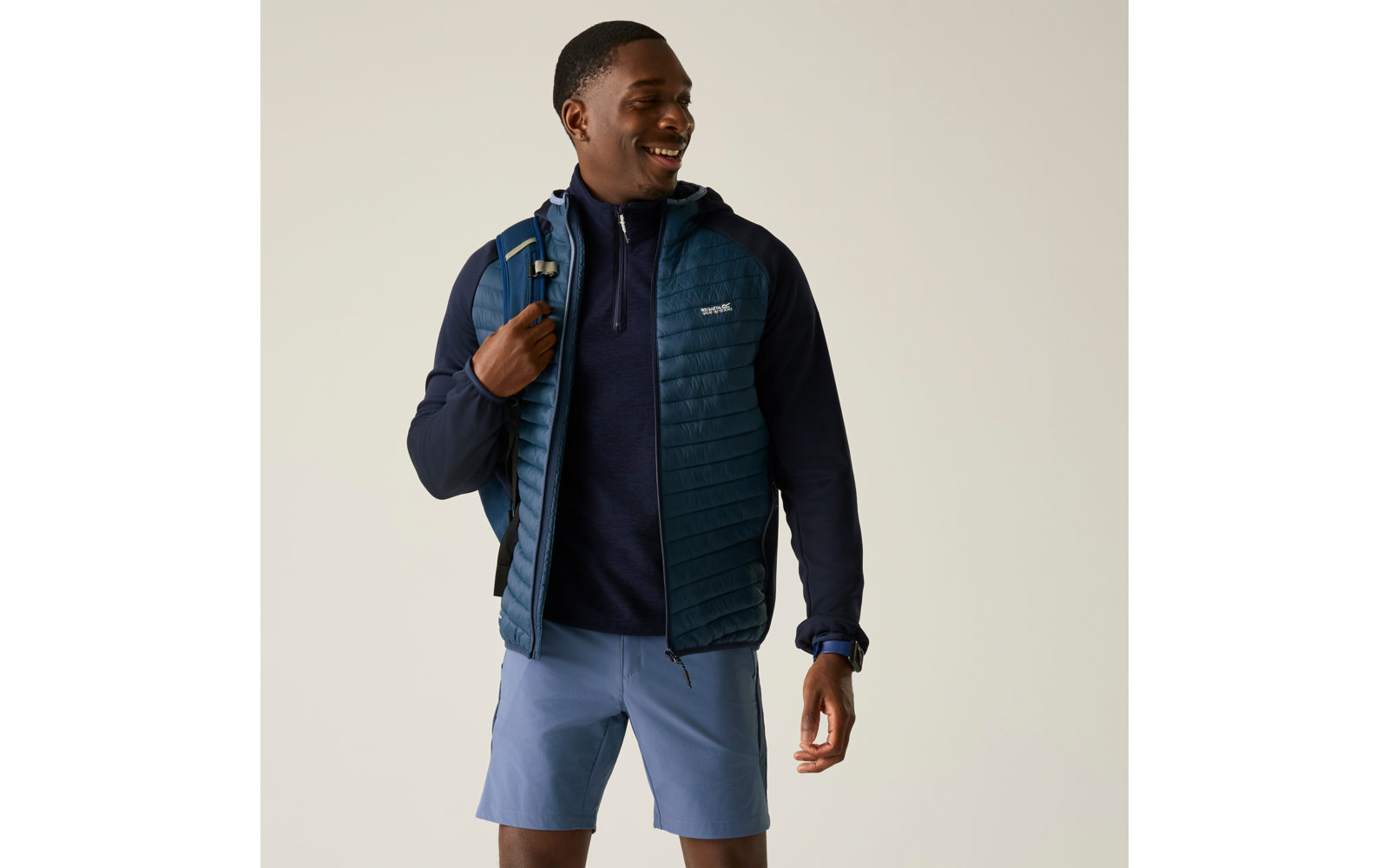 Person steht vor hellem Hintergrund, trägt dunkelblaue Steppweste über Pullover, hellblaue Shorts, Rucksack auf linker Schulter, schaut nach rechts und lächelt