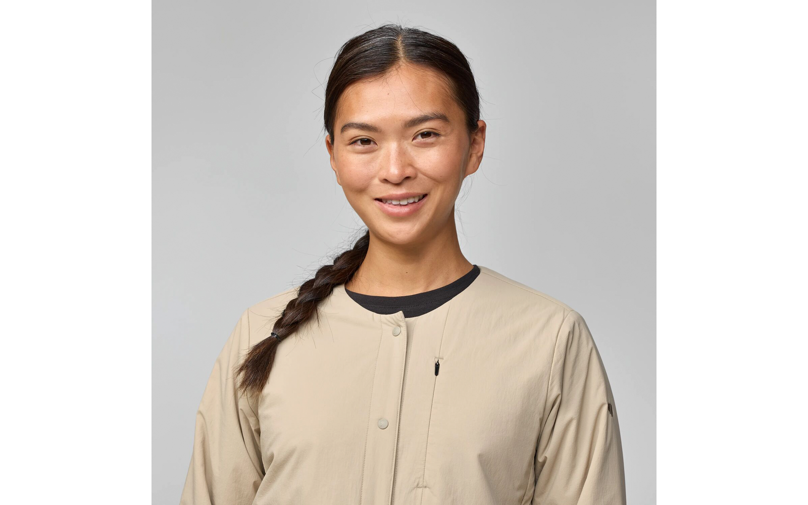 Person mit seitlichem Zopf, beige knopfbare Jacke und schwarzes Oberteil, leichtes Lächeln, hellgrauer Hintergrund