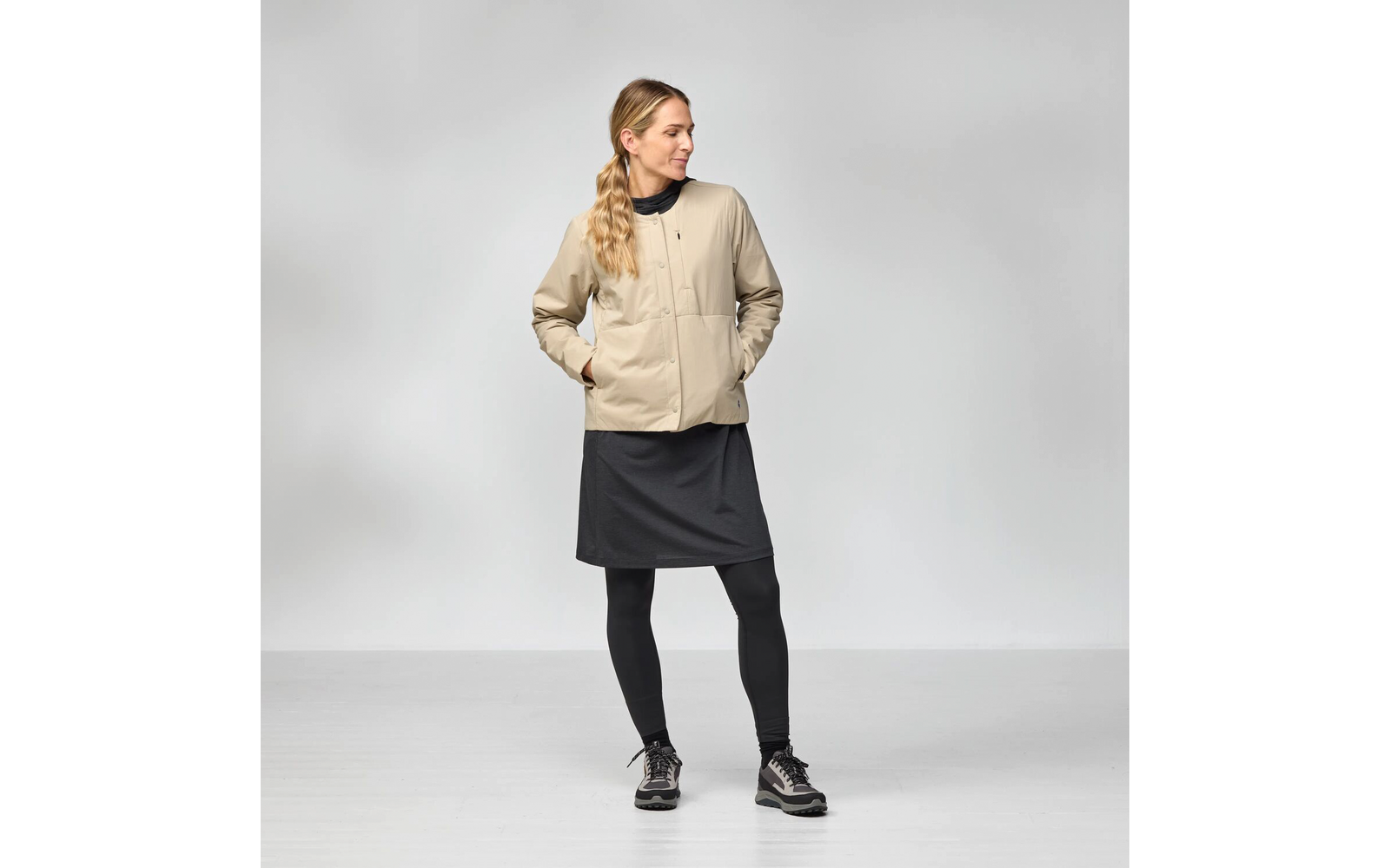 Person mit beiger Jacke, schwarzem Kleid, schwarzen Leggings und Schnürschuhen, Hände in den Taschen, Blick nach links, seitlicher Zopf, heller Hintergrund