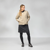Frau steht vor hellem Hintergrund, trägt beige Jacke, dunkles knielanges Kleid, schwarze Leggings und Sportschuhe, Hände in den Taschen, Blick nach rechts