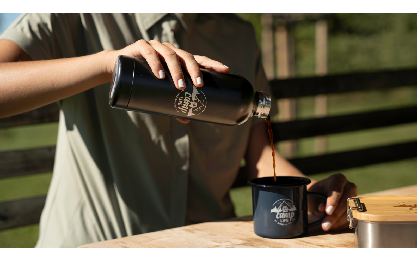 Handen van een persoon schenken donkere vloeistof uit een zwarte thermoskan in een donkerblauwe emaille mok op een houten tafel; houten schutting op de achtergrond