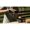 Mains d'une personne versant un liquide foncé d'une thermos noire dans une tasse émaillée bleu foncé sur une table en bois ; clôture en bois en arrière-plan