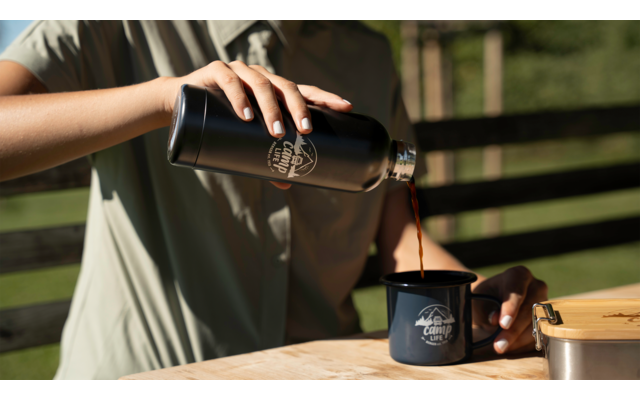 Mains d'une personne versant un liquide foncé d'une thermos noire dans une tasse émaillée bleu foncé sur une table en bois ; clôture en bois en arrière-plan