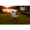 Klappstuhl auf Graswiese am Ufer eines Sees bei Sonnenuntergang, Bäume und Berge im Hintergrund, geöffneter Camper mit sichtbarem Innenraum rechts