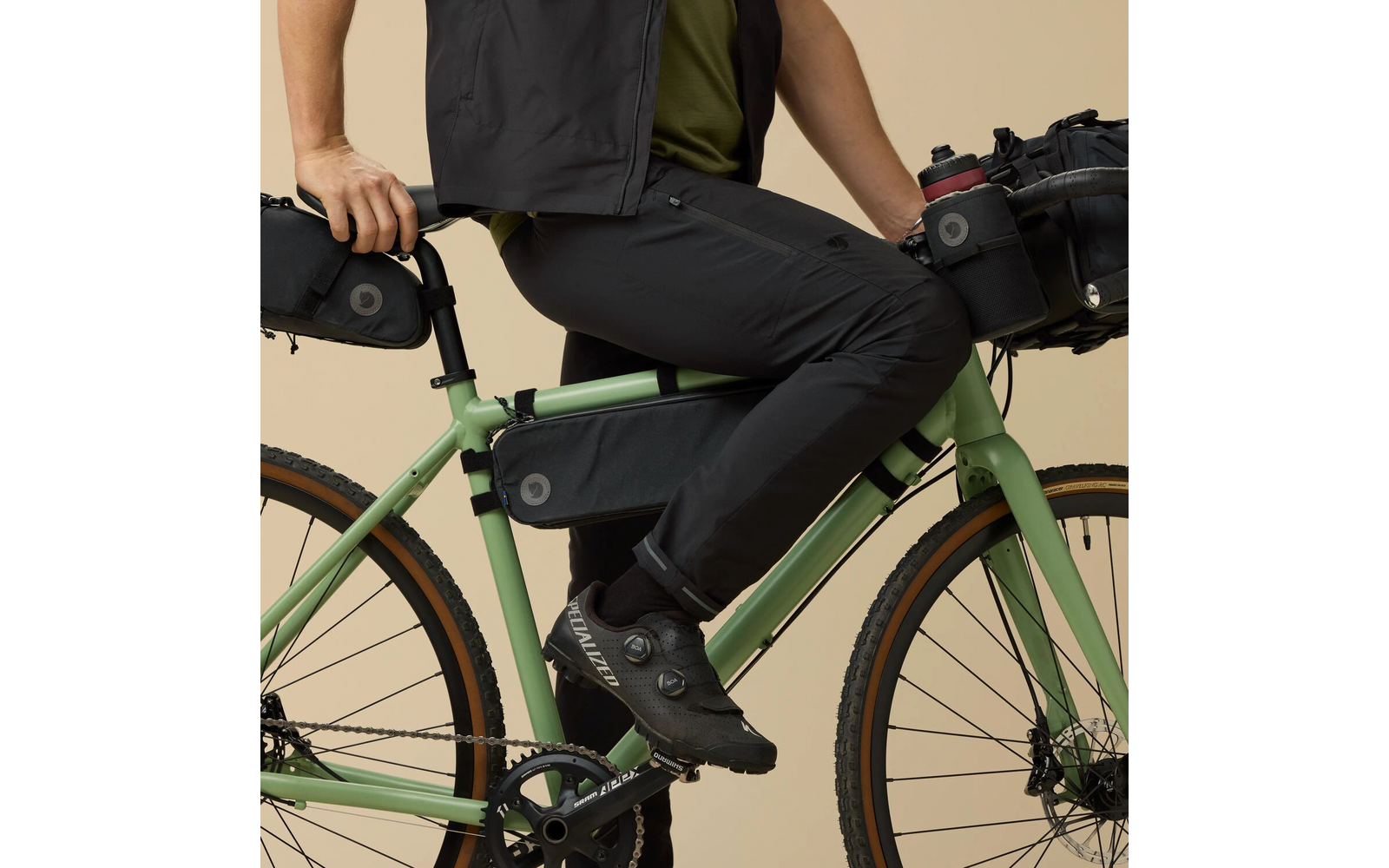 Persoon zit op een lichtgroene fiets. Zichtbaar: bovenlichaam tot onderbenen, olijfgroen T‑shirt, zwart vest en zwarte fietsbuis, zwarte fietsschoenen, hand op zadel of achtertas, zwarte frametas aan het onderbuis, zwarte achtertas, lichtbruine bandzijwanden, kettingblad en pedaal