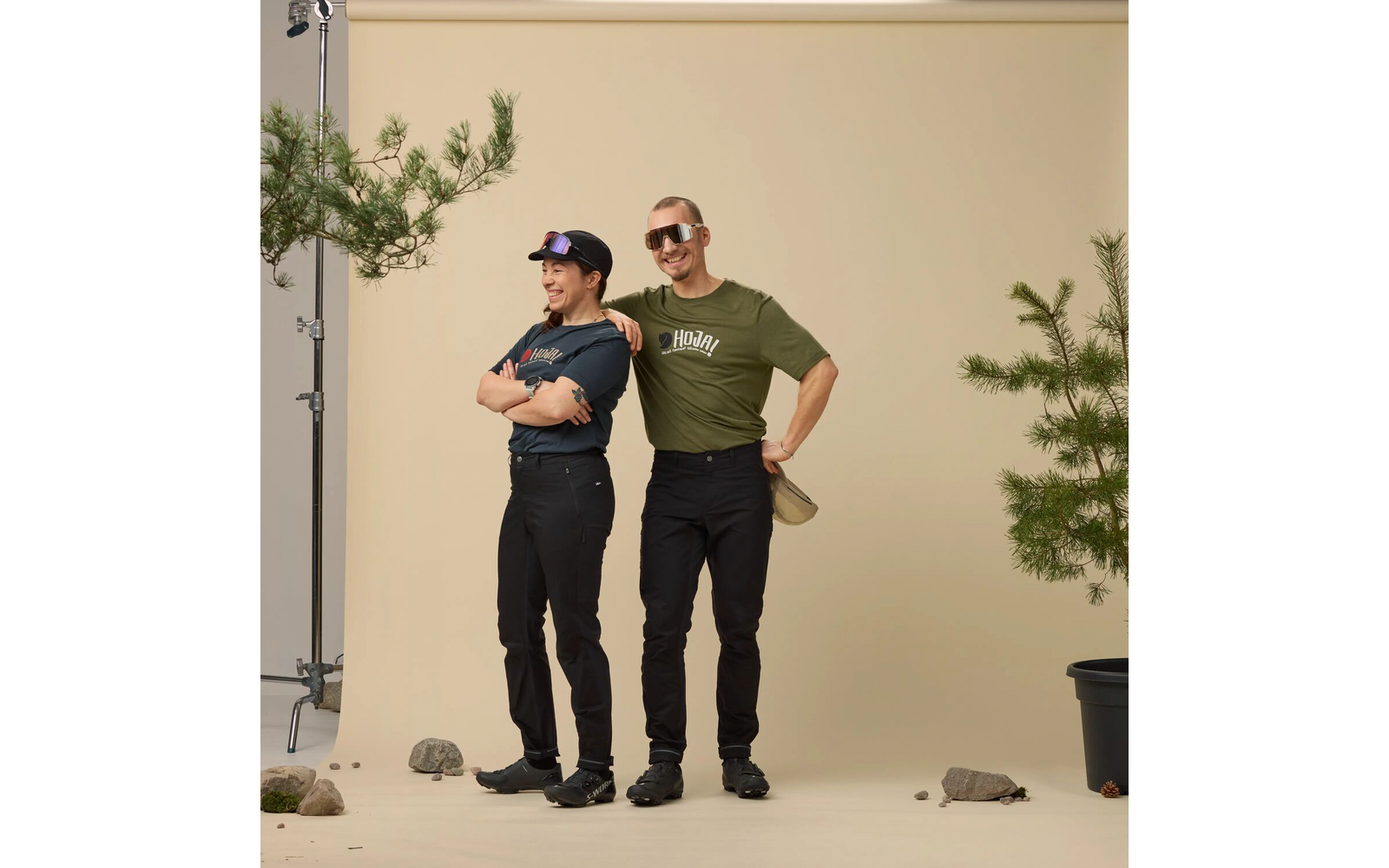 Twee personen staan voor een beige achtergrond; links met pet en armen over elkaar, rechts met zonnebril en groen T‑shirt en één hand achter de rug; beiden dragen zwarte broeken en schoenen; links een lampstatief, rechts een pot met een kleine den en stenen op de vloer