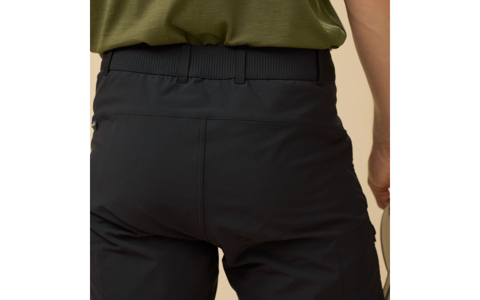 Rugaanzicht van de taille en heupen van een persoon met een groen T‑shirt, ingestopt in een zwarte broek met elastische taille en riemlussen; rechterhand houdt een beige voorwerp