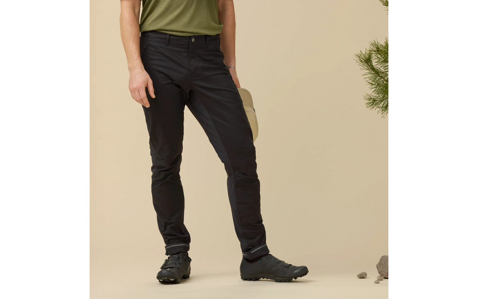 Onderlichaam van een persoon met een olijfgroen bovenstuk, donkere broek en zwarte wandelschoenen, in de rechterhand een lichtbeige pet; een dennentak en stenen op een beige achtergrond