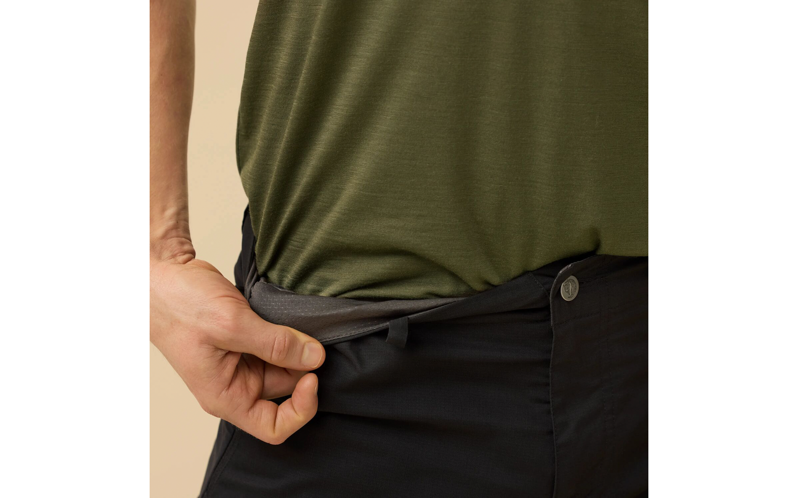 Close-up van een bovenlichaam met een olijfgroen shirt, een hand trekt de tailleband van een donkere broek naar buiten, zichtbare knoop