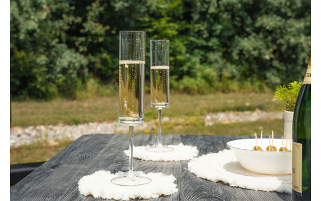 Deux flûtes de champagne remplies sur des sous-verres blancs sur une table en bois foncé, à côté d'un bol d'olives et du bord d'une bouteille ; herbe et buissons en arrière-plan