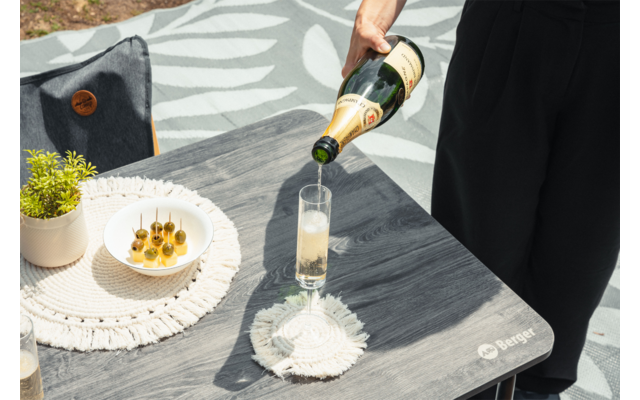Une main verse une boisson claire et pétillante d'une bouteille verte dans une flûte étroite sur une table en bois grise ; sur la table se trouvent deux dessous-de-verre ronds tissés, un bol avec de petites bouchées vertes sur des piques et un petit pot de plante