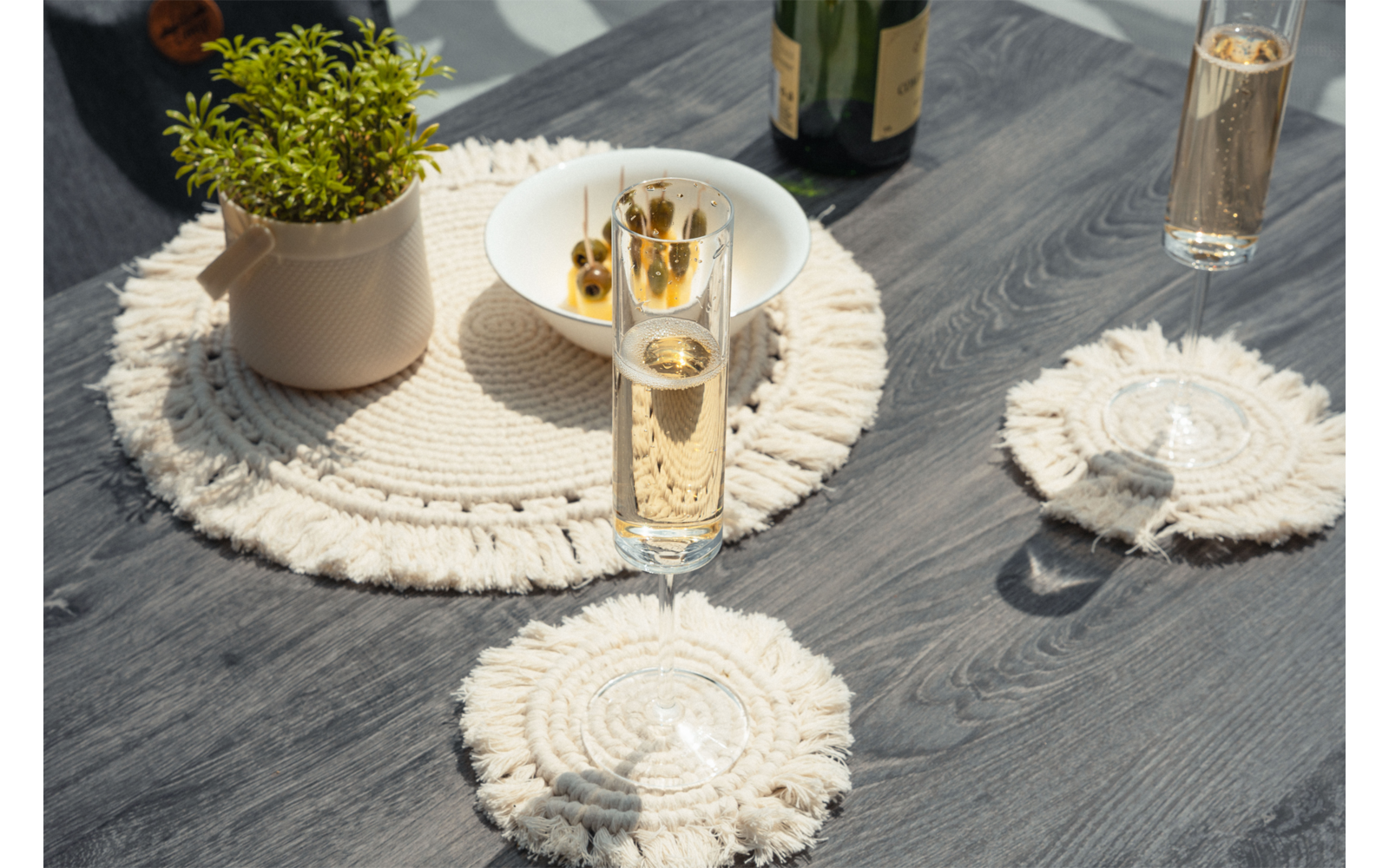 Table en bois foncé avec des sets ronds en macramé crème, deux flûtes contenant une boisson dorée, une bouteille, un bol blanc d'olives et un petit pot avec une plante verte