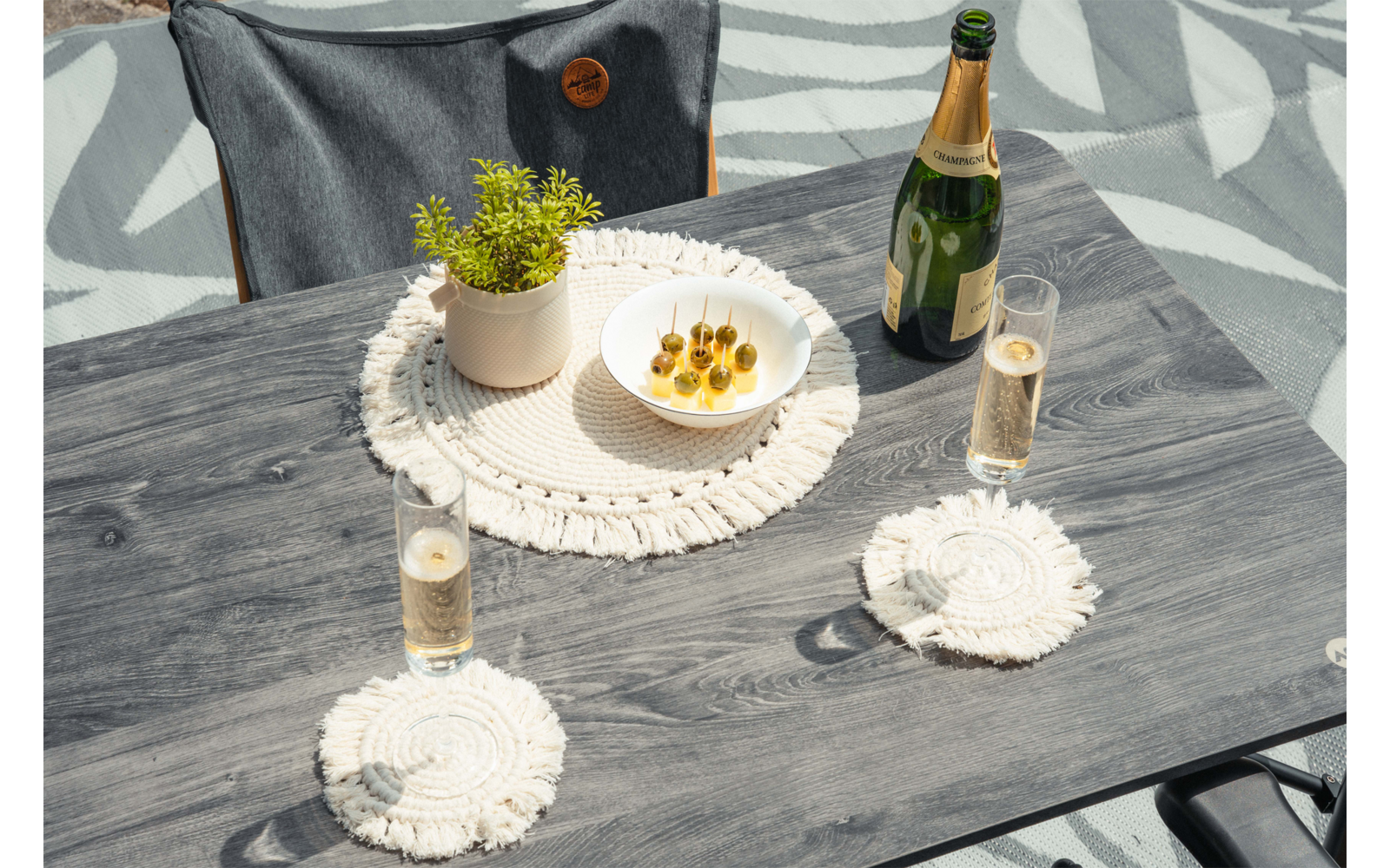 Table grise avec une bouteille à capsule dorée, deux verres remplis, un bol blanc avec trois brochettes, une petite plante en pot et des sets de table crochetés ; chaise grise en arrière-plan