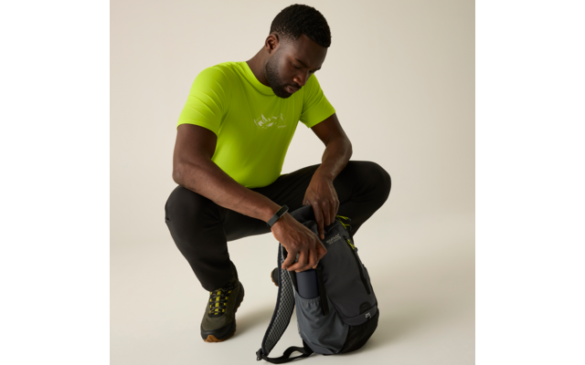 Persoon hurkt, draagt een neon groen T-shirt, zwarte broek en wandelschoenen, stopt een smartphone in de zijzak van een grijze rugzak voor een lichte achtergrond