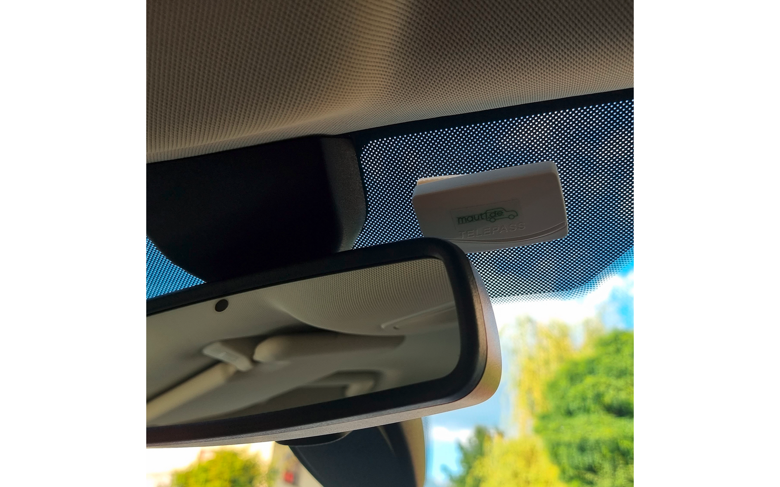 Innenraum eines Autos mit Rückspiegel; oberhalb des Spiegels an der Windschutzscheibe ein kleines weißes Kästchen; punktierte Folie am Glas; draußen Bäume und blauer Himmel