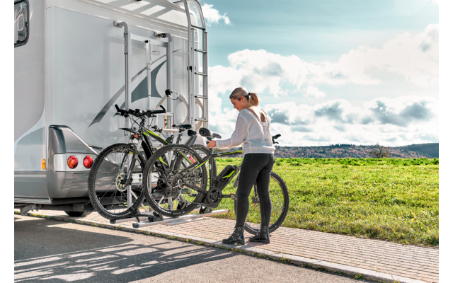 Femme fixant un vélo au porte-vélos arrière d’un camping-car blanc sur une aire pavée à côté d’une prairie sous un ciel nuageux