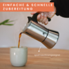Mano vierte café de una cafetera moka de acero inoxidable en una taza de cerámica sobre una superficie de madera, vapor visible; banda naranja con el texto "EINFACHE & SCHNELLE ZUBEREITUNG" y flecha con "spülmaschinengeeignet"