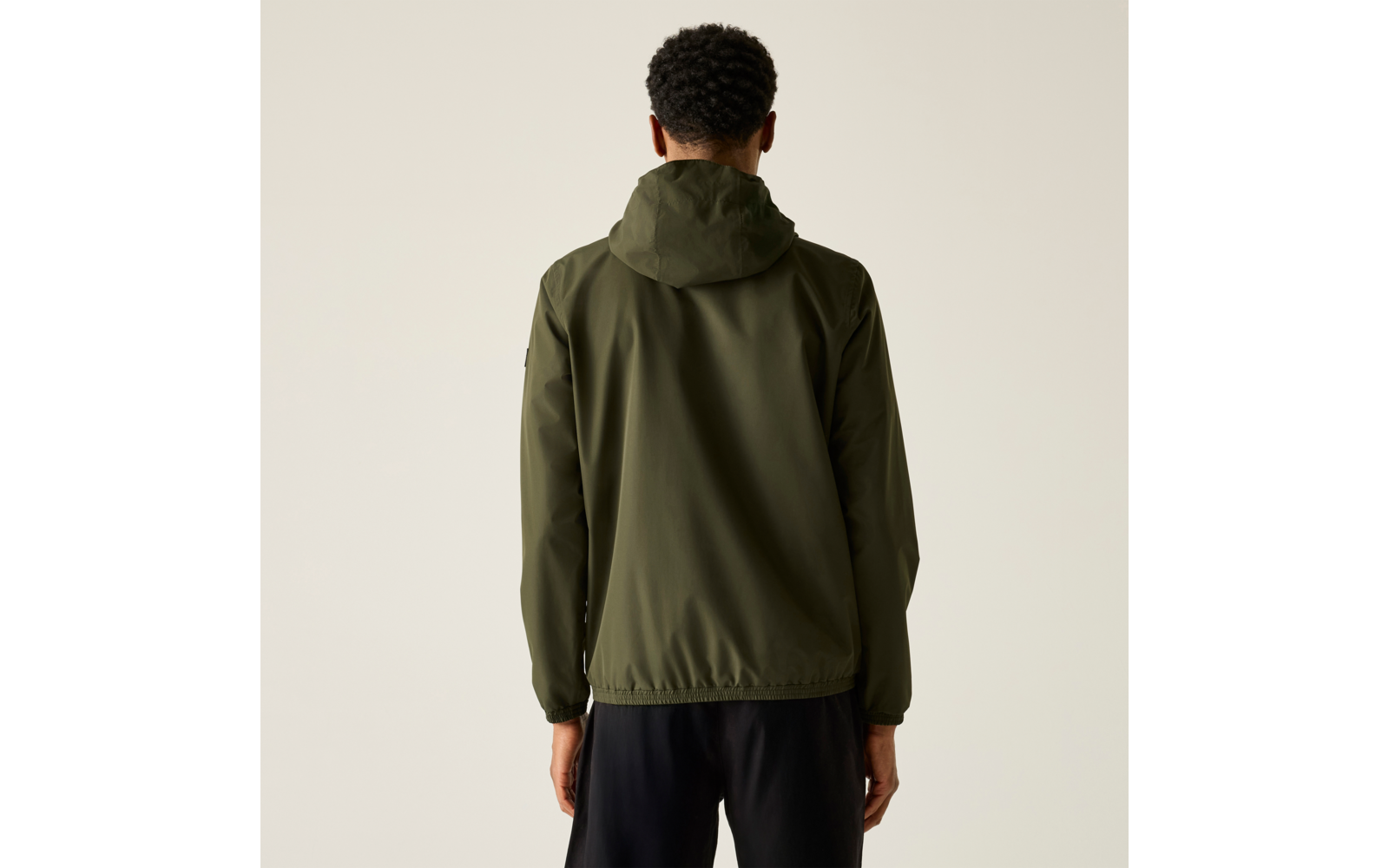 Person von hinten mit kurzen dunklen lockigen Haaren, trägt olivgrüne Kapuzenjacke und schwarze Hose vor hellem beigem Hintergrund