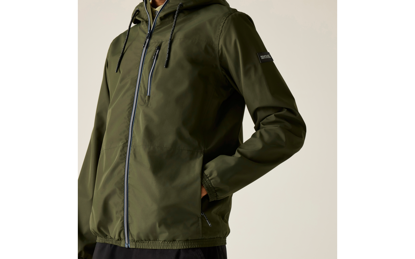 Oberkörper einer Person in olivgrüner Kapuzenjacke mit zentralem Reißverschluss, zwei vertikalen Brustreißverschlüssen, Kordeln und einer Hand in der seitlichen Tasche