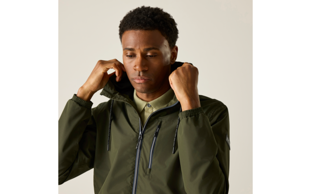 Mann mit kurzen, lockigen Haaren, trägt olivgrüne Kapuzenjacke mit Reißverschluss und beige Poloshirt, hält die Kapuze mit beiden Händen, heller einfarbiger Hintergrund