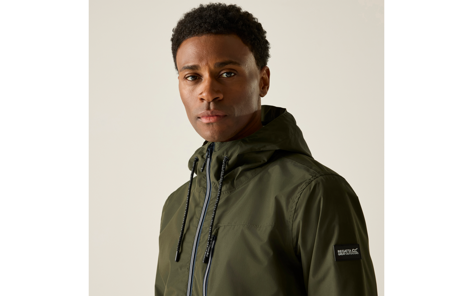 Porträt einer Person mit kurzem, lockigem, dunklem Haar, die eine olivgrüne Kapuzenjacke mit Reißverschluss und Kordeln trägt; heller einfarbiger Hintergrund