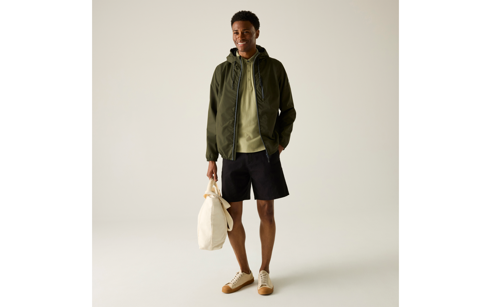 Person steht vor hellem neutralem Hintergrund, trägt olivgrüne Jacke über hellgrünem Kapuzenpullover, schwarze Shorts, beige Sneaker und hält eine helle Stofftasche