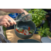 Handen met een zilveren ring houden twee gesleufde spatels boven een grijze kom met gesneden rode paprika, uienringen en groene groentestukjes; op de achtergrond een kruidentje op een houten tafelblad