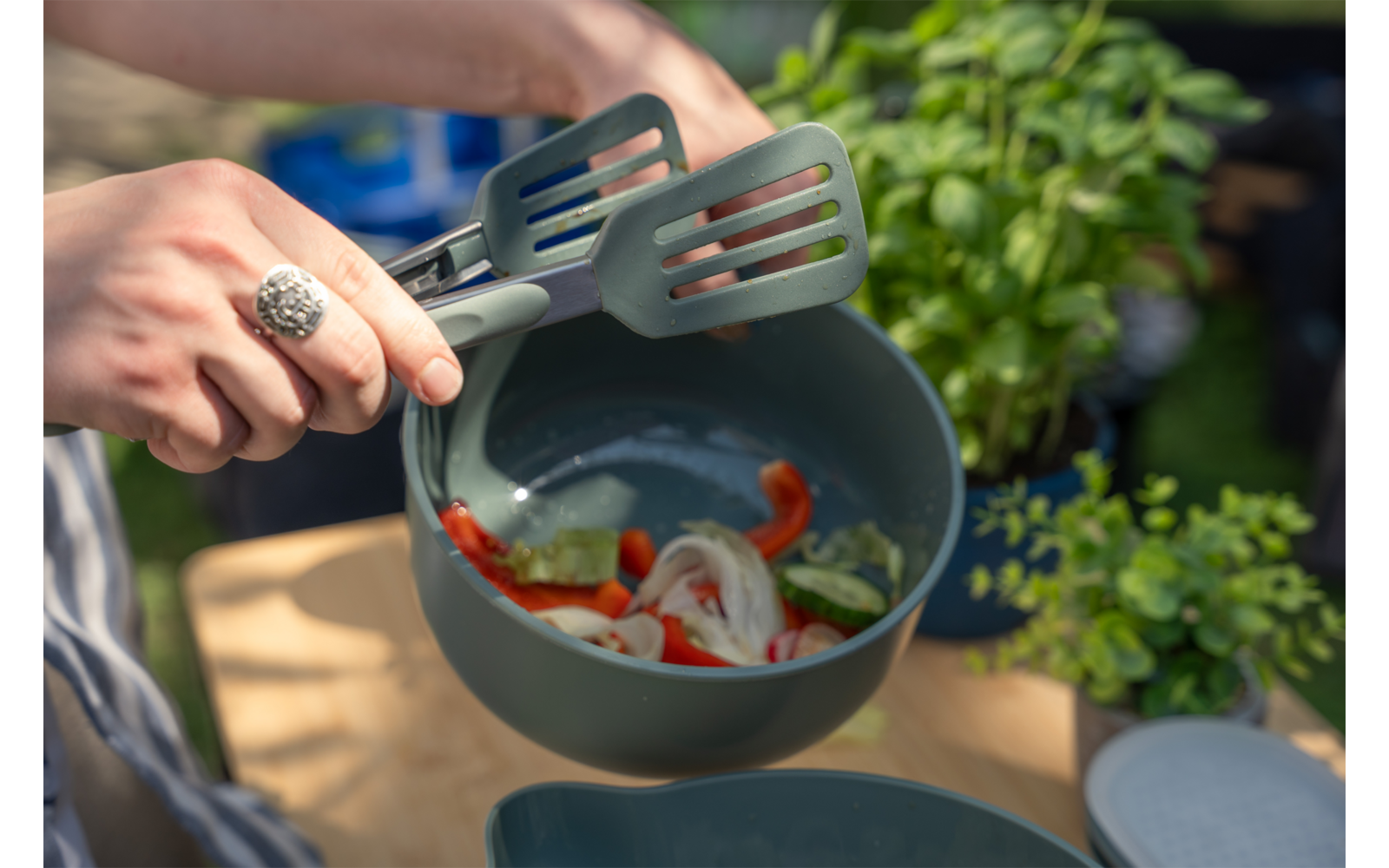 Handen met een zilveren ring houden twee gesleufde spatels boven een grijze kom met gesneden rode paprika, uienringen en groene groentestukjes; op de achtergrond een kruidentje op een houten tafelblad